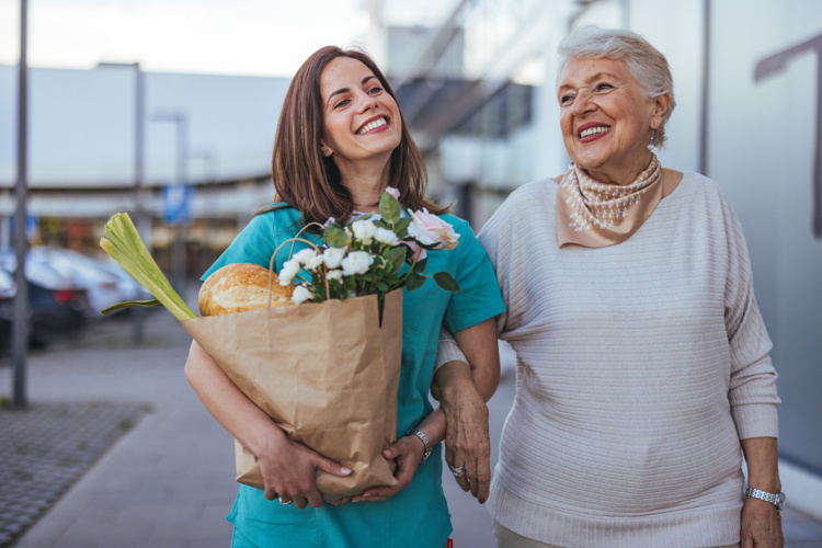 Two women are smiling. One is carrying groceries in a paper bag. 