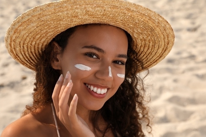 woman in wide rim, straw hat applying sun screen to her face as she smiles at the camera.