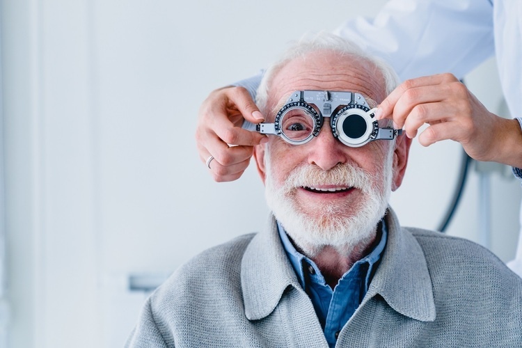 Older white male facing the camera, smiling with instrument partially obstructing his face. Doctor stands behind while holding edges of instrument to test vision.