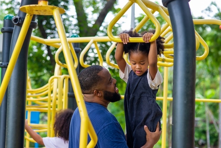 child holding onto monkeybars with  man supporting her legs