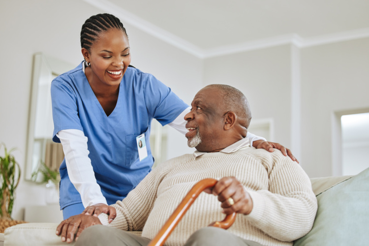 Woman in scrubs is leaning towards older adult male who is seated and holding a cane